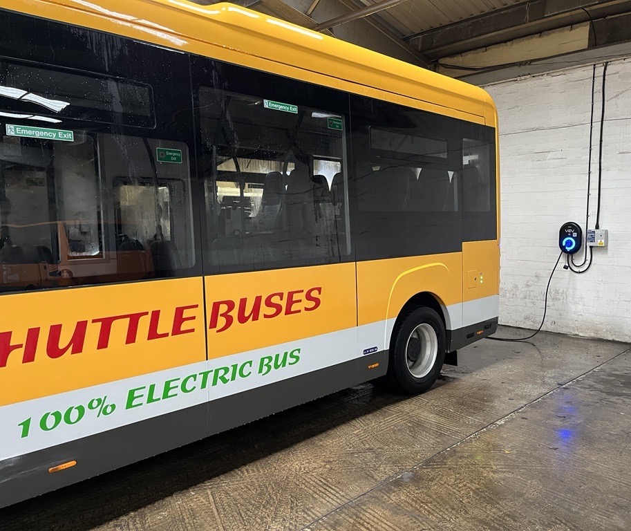 A yellow single-level electric bus with red Shuttle Buses logo plugged into a black wall charger inside a depot building with grey walls