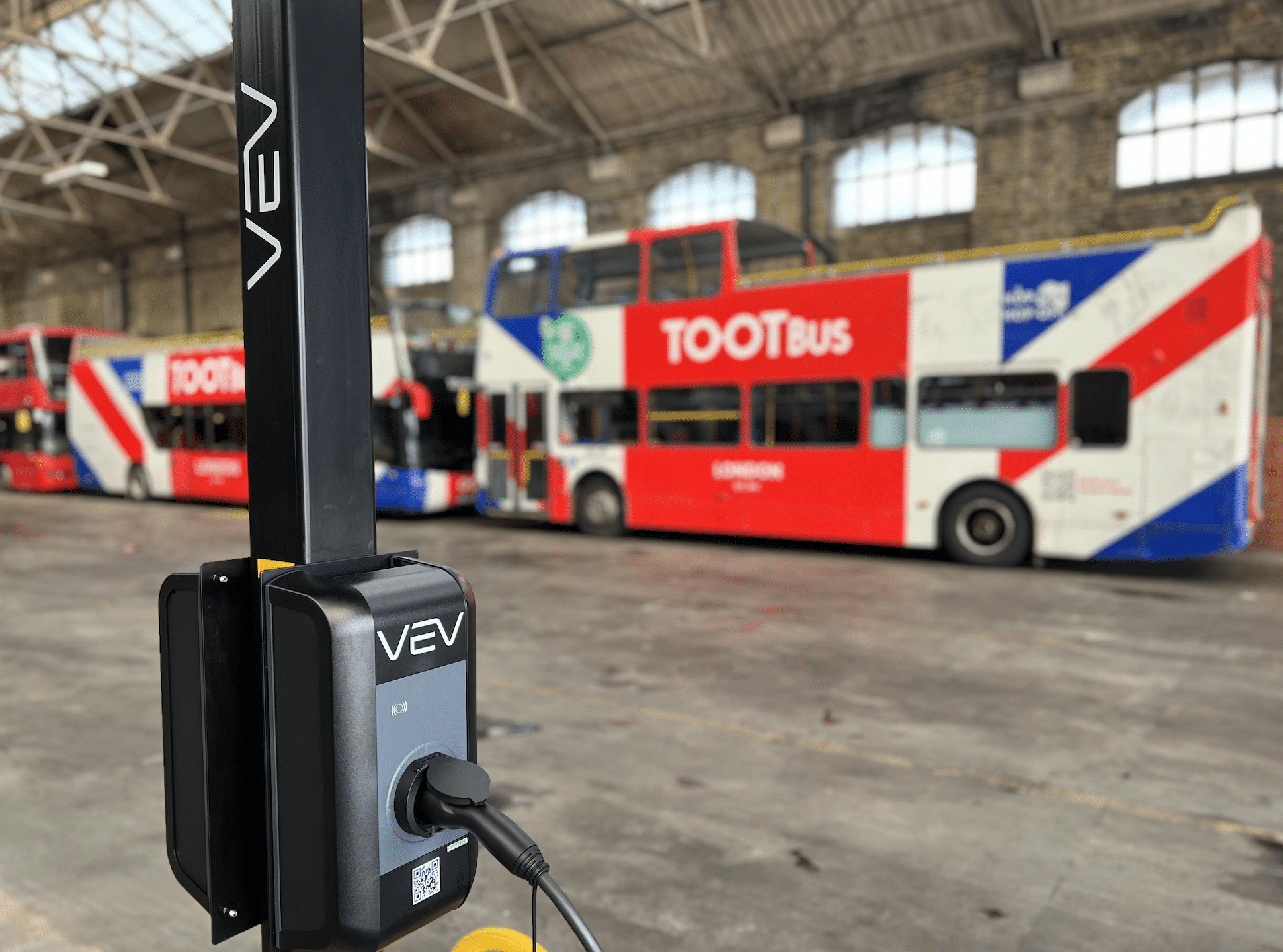 A Tootbus branded electric open-top tour bus with Union Jack colouring in a depot, behind a black, VEV-branded charge point on a VEV branded pillar