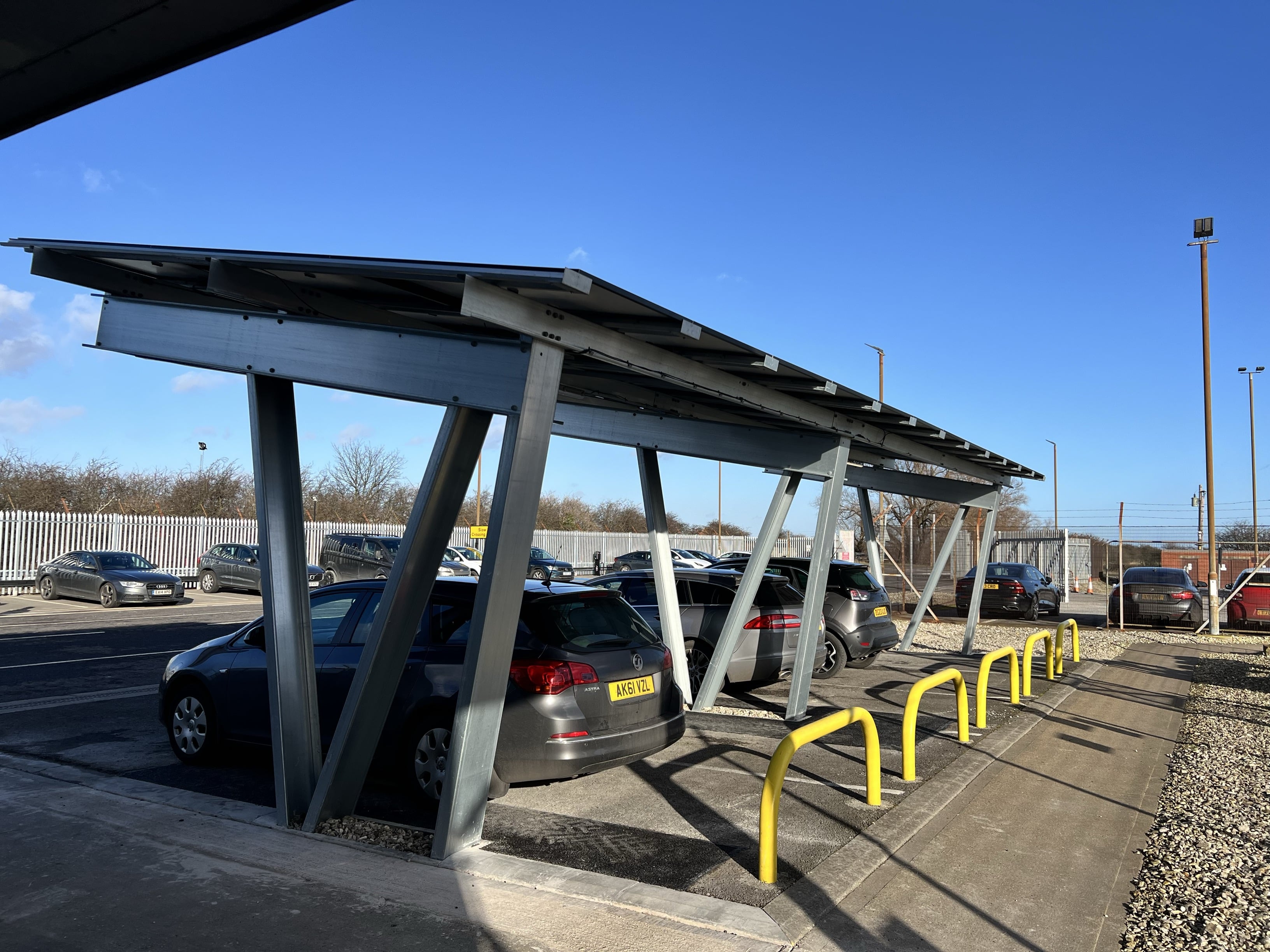 Three cars parked beneath a solar canopy structure which is generating power to charge vehicles. Background of industrial area carpark with clear blue sky