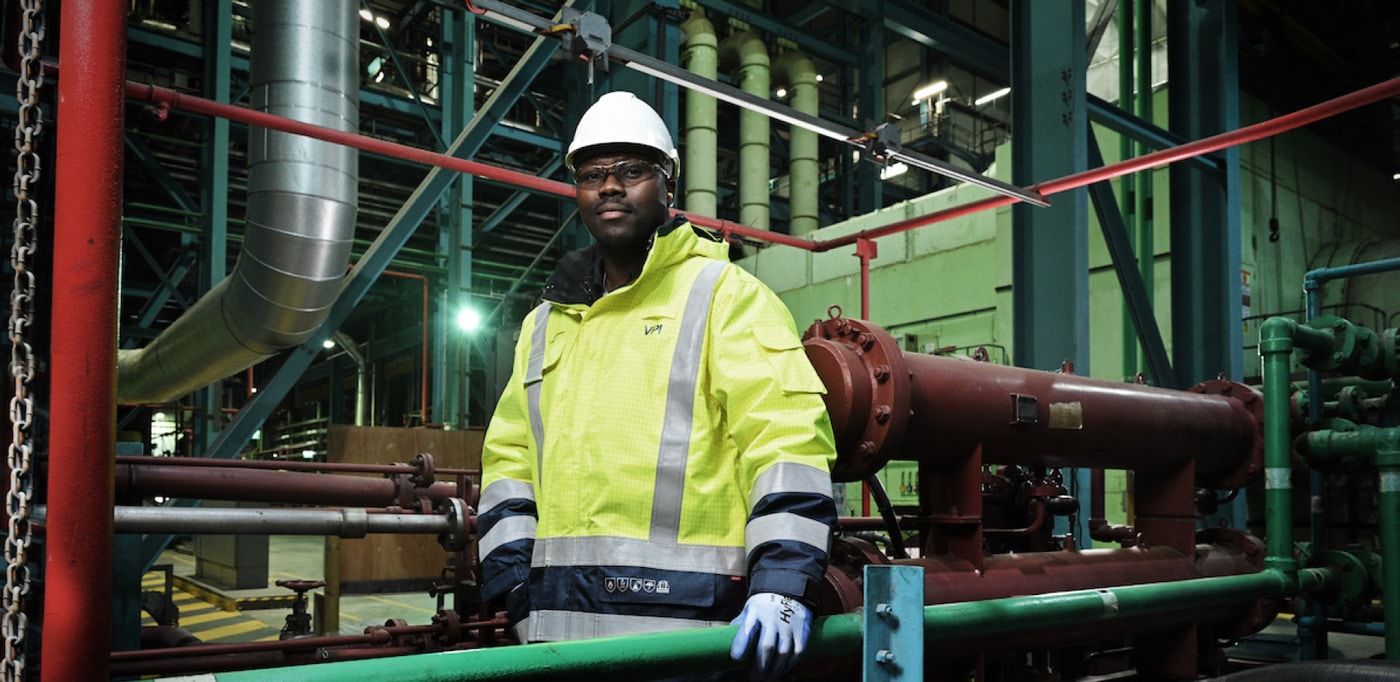 Factory operative in yellow hi-vis and white protective headgear looks at the camera in a factory, backdrop of pipes and metal frameworks