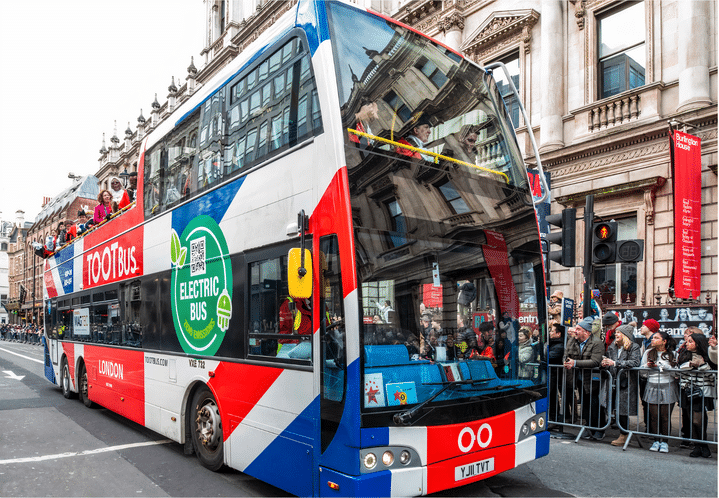 Many passengers on a Tootbus branded open-top tour bus with large green 'Electric Bus' sticker and Union Jack patterning on a Central London Street, with grand buildings and lots of tourists